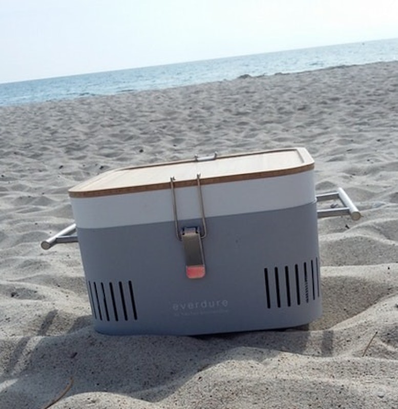 Der Everdure Cube in grau steht am Strand, im Hintergrund das Meer.