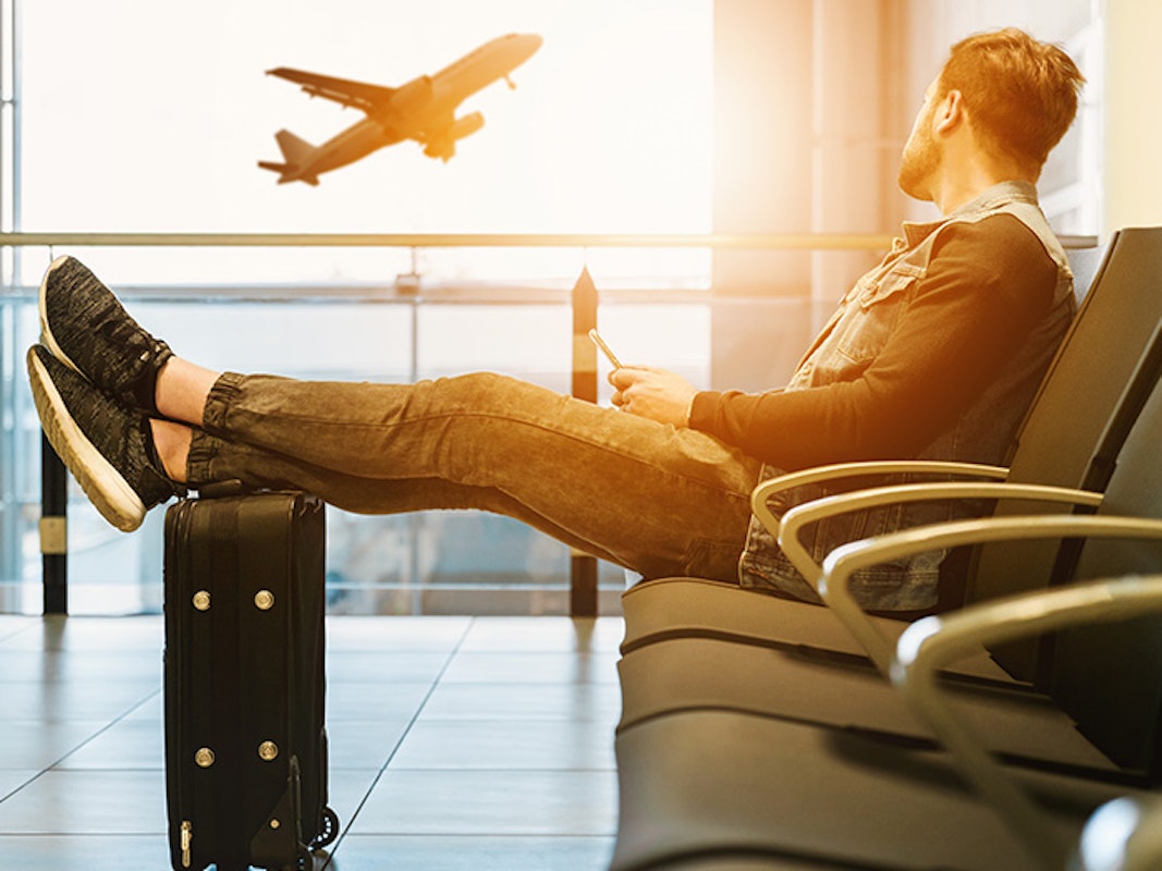 Ein Mann sitzt im Flughafen und schaut gebannt zu einem Flugzeug, das gerade abhebt. Seine Augen verfolgen das Flugzeug am Himmel, während er gleichzeitig entspannt mit seinen Füßen auf seinem Koffer ruht. 