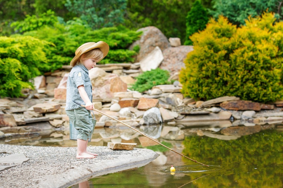 Kleiner Junge mit Strohhut angelt im Teich.