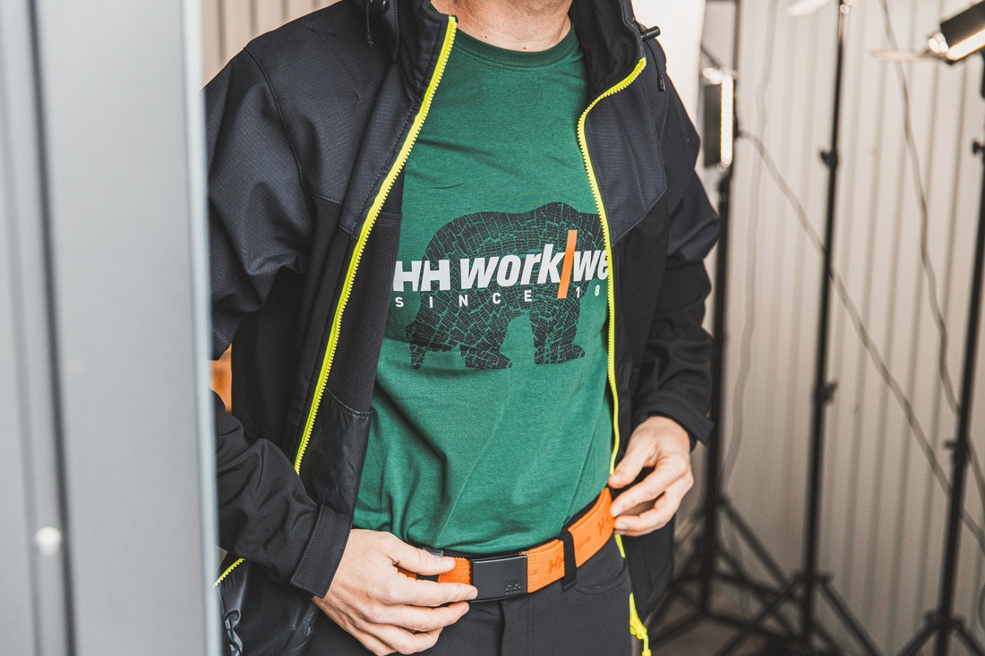 Arbeitskleider wie orangene Gürtel, grünes Arbeitsshirt  und schwarze Arbeitshose und Softshelljacke der Marke Helly Hansen