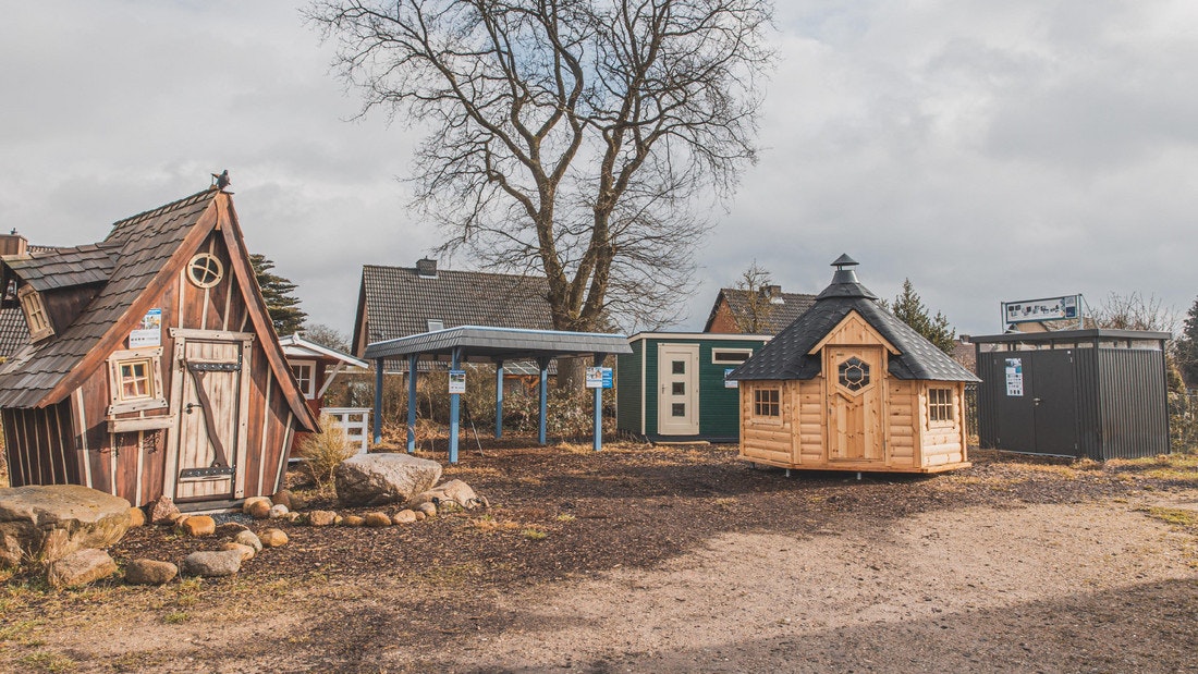 Ausstellung Reinbek Panorama Gartenhausausstellung mit mehreren Holzhütten, Metallgartenhäusern und einem blauen Carport