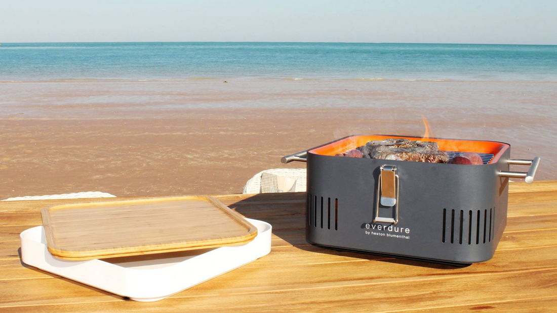 Everdure Cube in Schwarz steht direkt am Strand auf einem Holztisch, der Strand und das Meer sind im Hintergrund